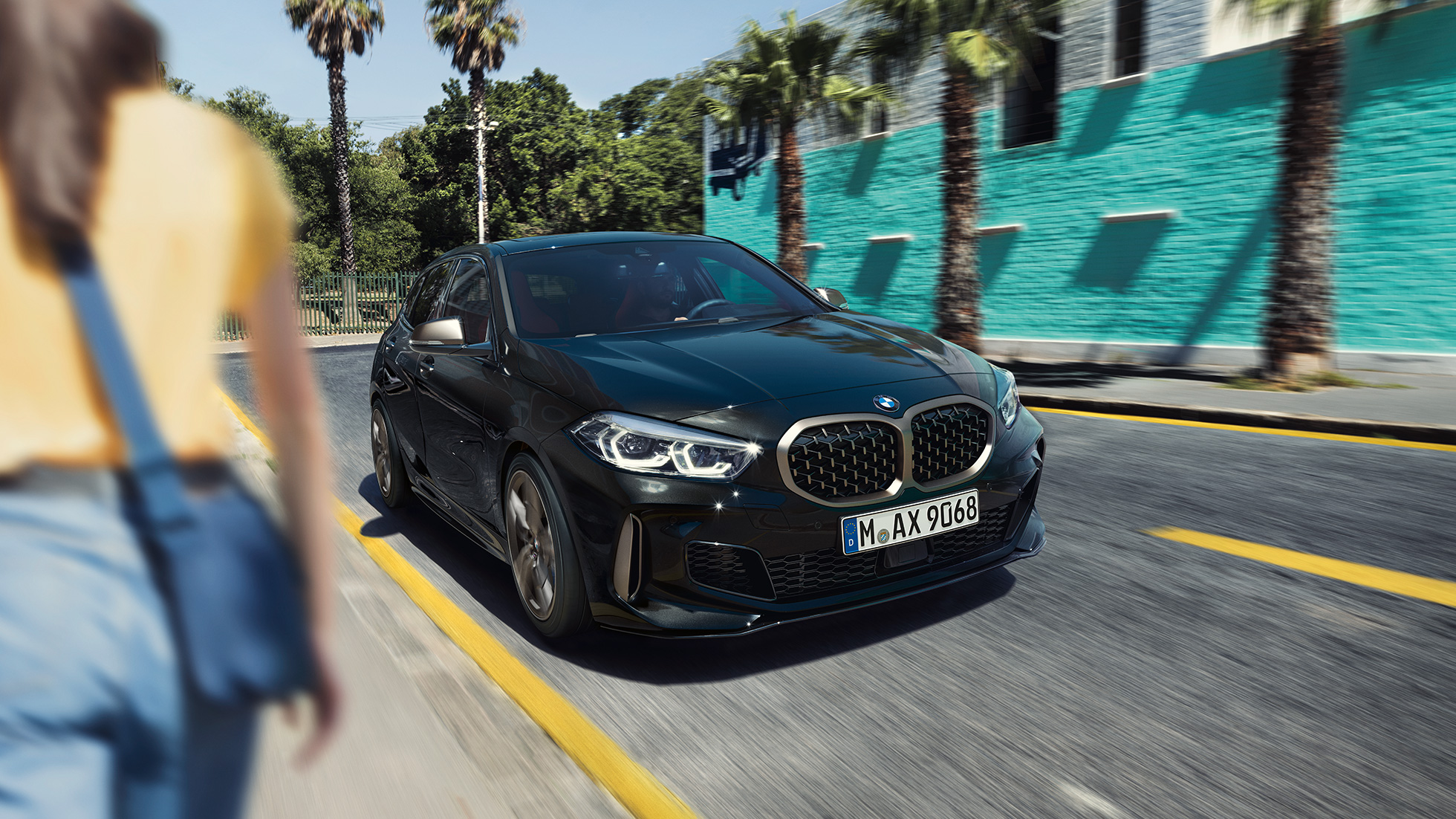 BMW M135i xDrive, vista frontal de tres cuartos. BMW M135i xDrive en Black Sapphire metallic; vista frontal de tres cuartos en movimiento, con una mujer en primer plano.