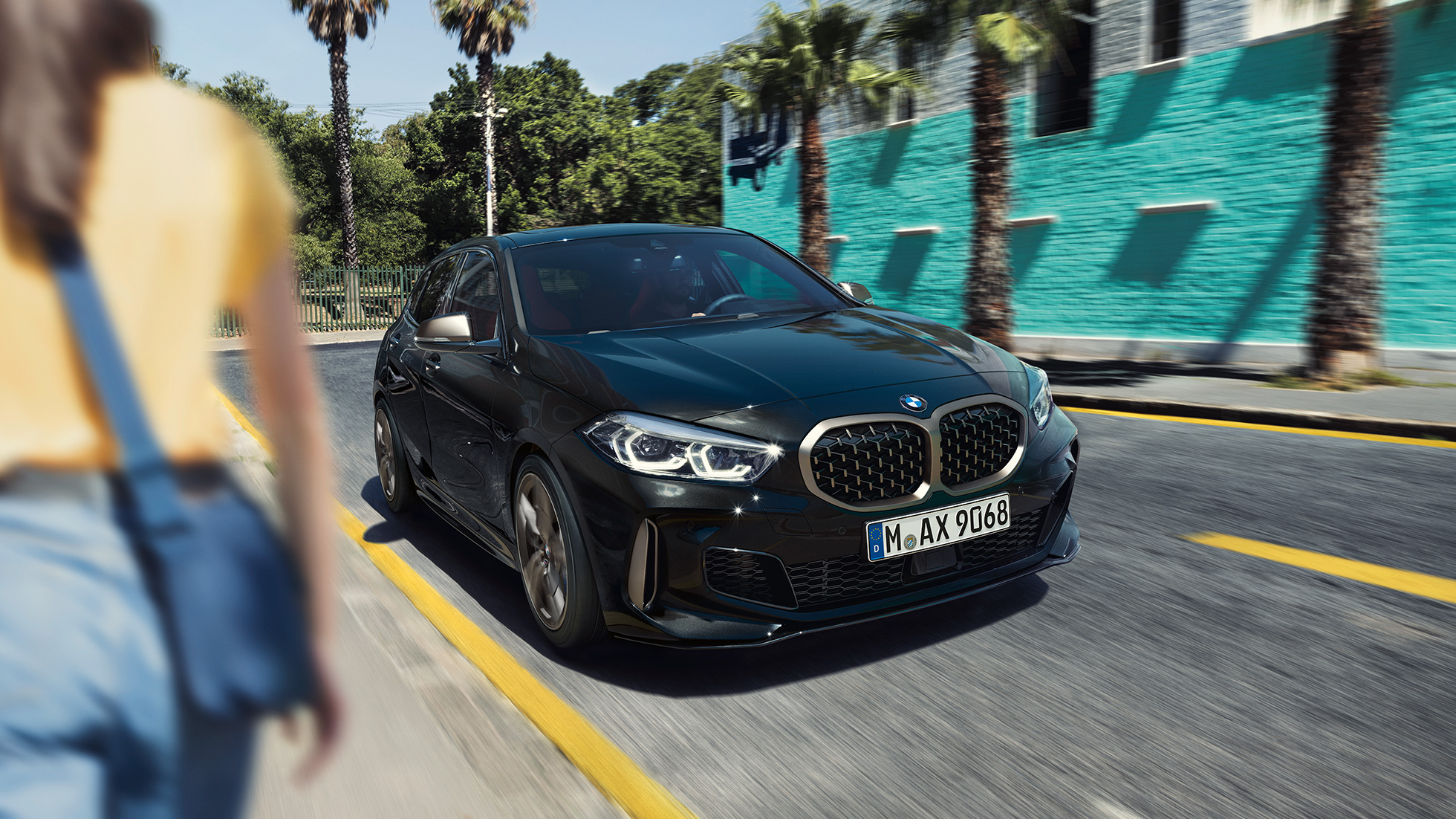 BMW M135i xDrive, vista frontal de tres cuartos. BMW M135i xDrive en Black Sapphire metallic; vista frontal de tres cuartos en movimiento, con una mujer en primer plano.